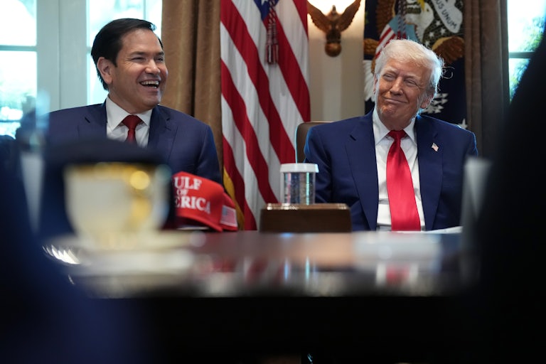 Donald Trump and Secretary of State Marco Rubio laugh while at a Cabinet meeting. A red "Gulf of America" cap is on the table in front of them.