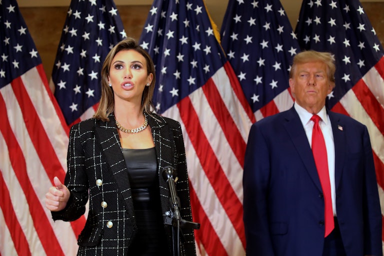 Alina Habba speaks in front of a mic, while Donald Trump stares at her. A row of U.S. flags are in the background.
