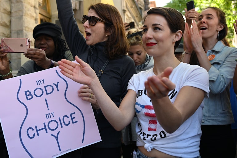 Two women clap and cheer in a crowd of protesters. One holds a sign that reads "My Body My Choice" and has the silhouette of a woman's body.