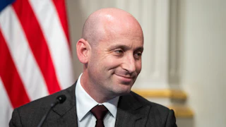 Stephen Miller is sitting wearing a dark suit and dark tie, with his head turned to the side with a smirk on his face.