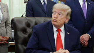 Donald Trump puckers his lips while sitting with his hands folded on his desk in the Oval Office