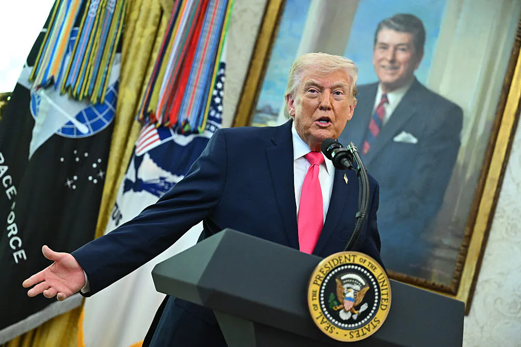 Donald Trump gestures with one hand while standing at a podium, in front of a portrait of Ronald Reagan.