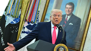 Donald Trump gestures with one hand while standing at a podium, in front of a portrait of Ronald Reagan.