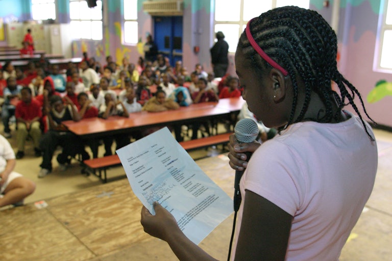 A Black girl in elementary school holds a mic and reads off a paper on stage.