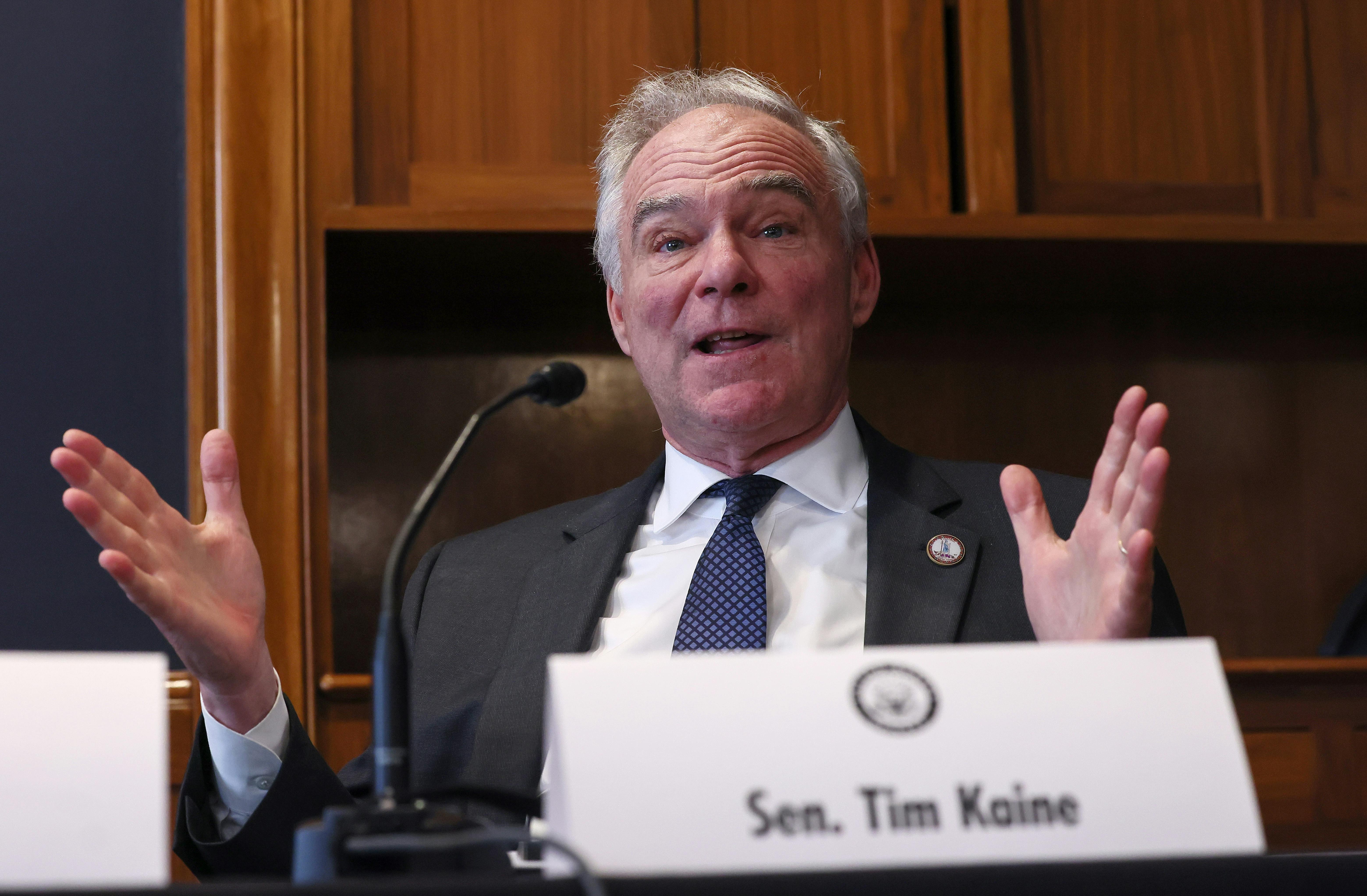 Senator Tim Kaine leans back in his chair and splays his hands out while speaking. A paper nameplate is on the table in front of him.
