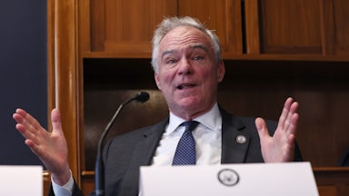 Senator Tim Kaine leans back in his chair and splays his hands out while speaking. A paper nameplate is on the table in front of him.