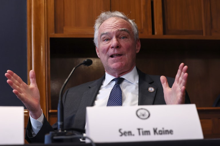 Senator Tim Kaine leans back in his chair and splays his hands out while speaking. A paper nameplate is on the table in front of him.