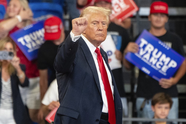 Donald Trump raises a fist as if in victory at a campaign rally. Others in the background hold Trump Vance signs.