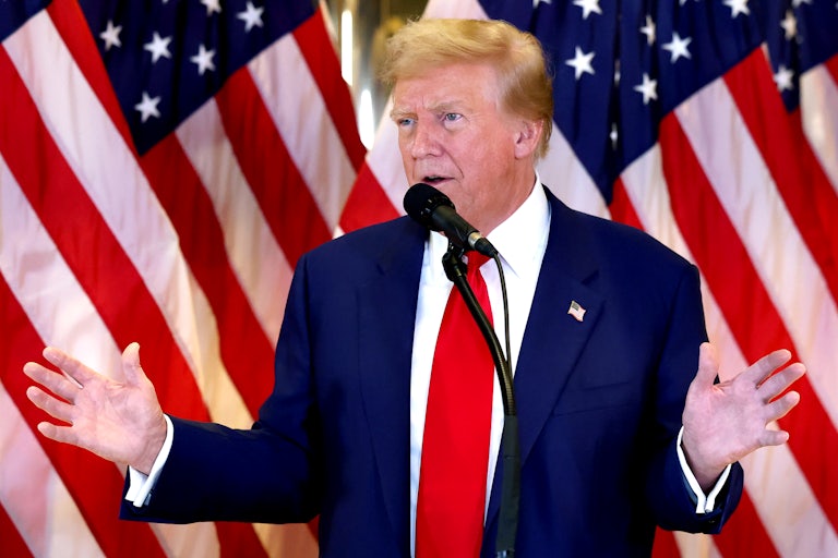 Donald Trump stands in front of U.S. flags speaking before a mic, hands splayed out