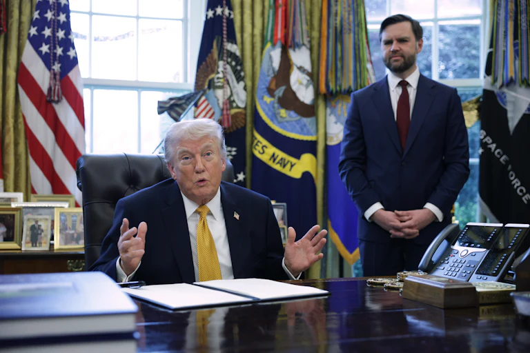 Donald Trump gestures and speaks while sitting at his desk in the Oval Office. JD Vance stands behind him.