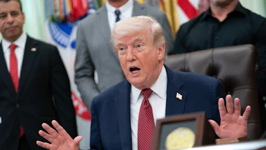 Donald Trump gestures with both hands and speaks while sitting at his desk in the Oval Office
