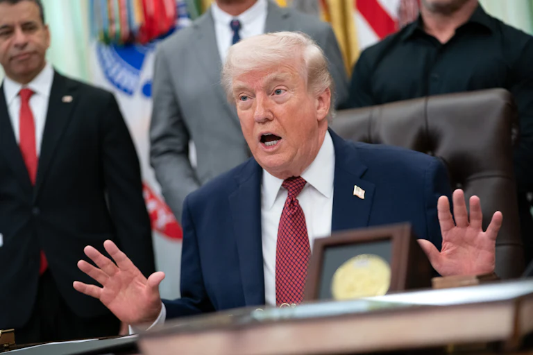 Donald Trump gestures with both hands and speaks while sitting at his desk in the Oval Office