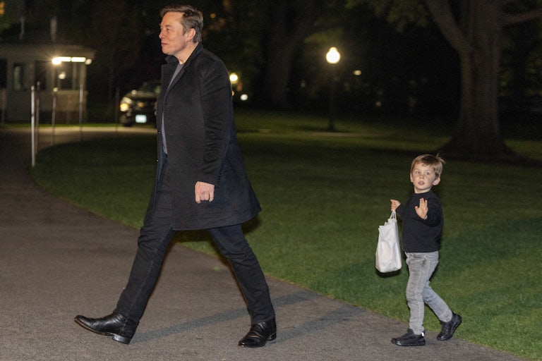 Elon Musk walks on the White House lawn at night. His young child X Æ walks behind him holding a bag in his hands and waving to the camera.