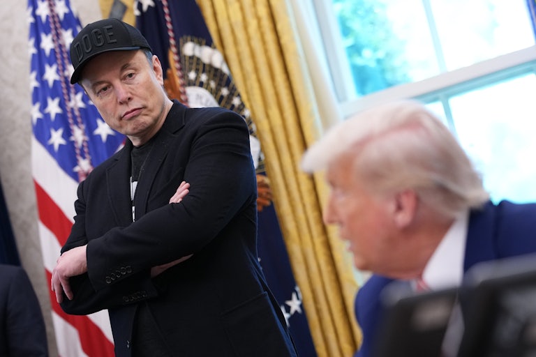 Elon Musk crosses his arms and looks down at Donald Trump, who is sitting at his desk in the Oval Office of the White House.