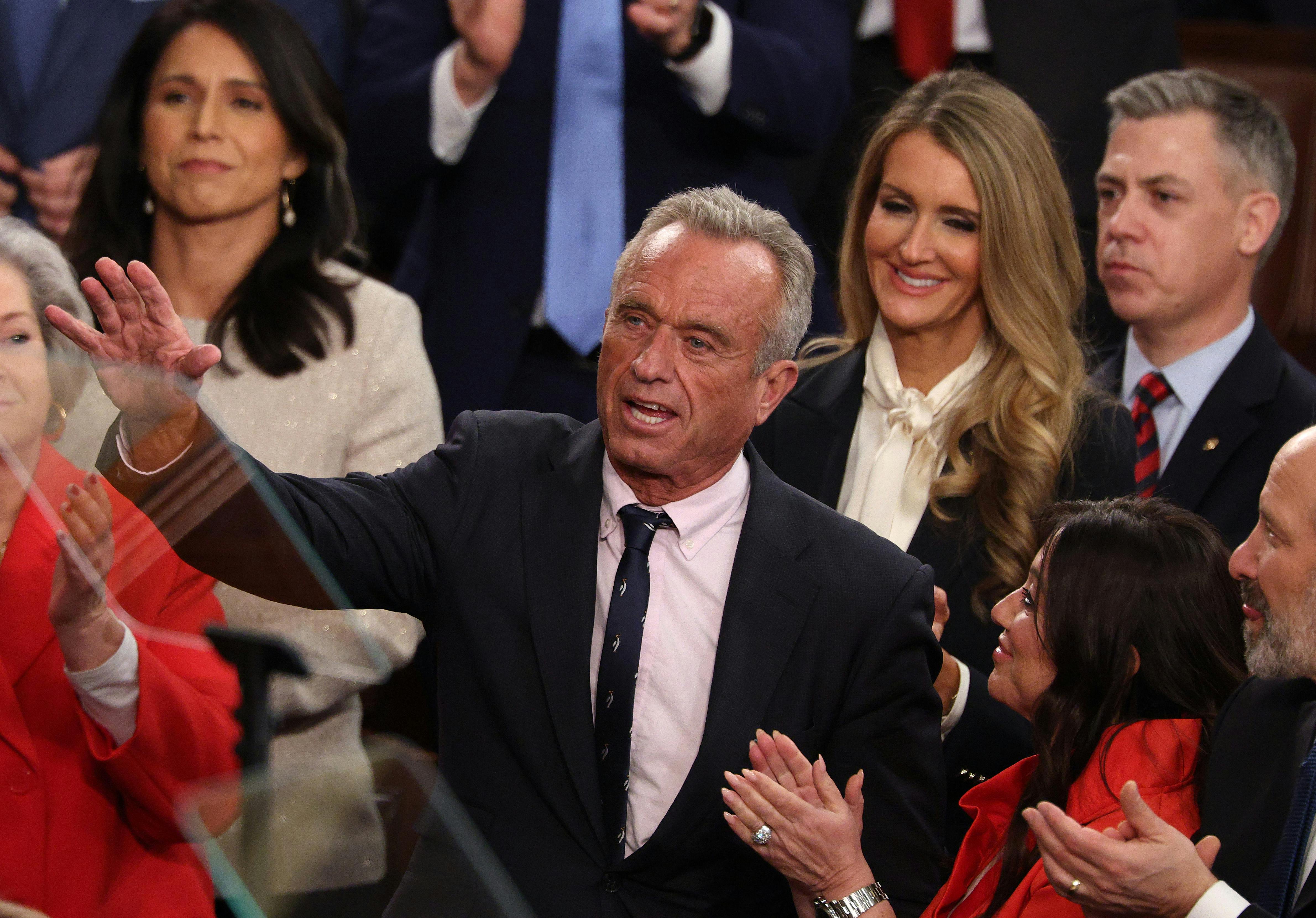 RFK Jr. smiles while raising a hand as people around him smile and clap.