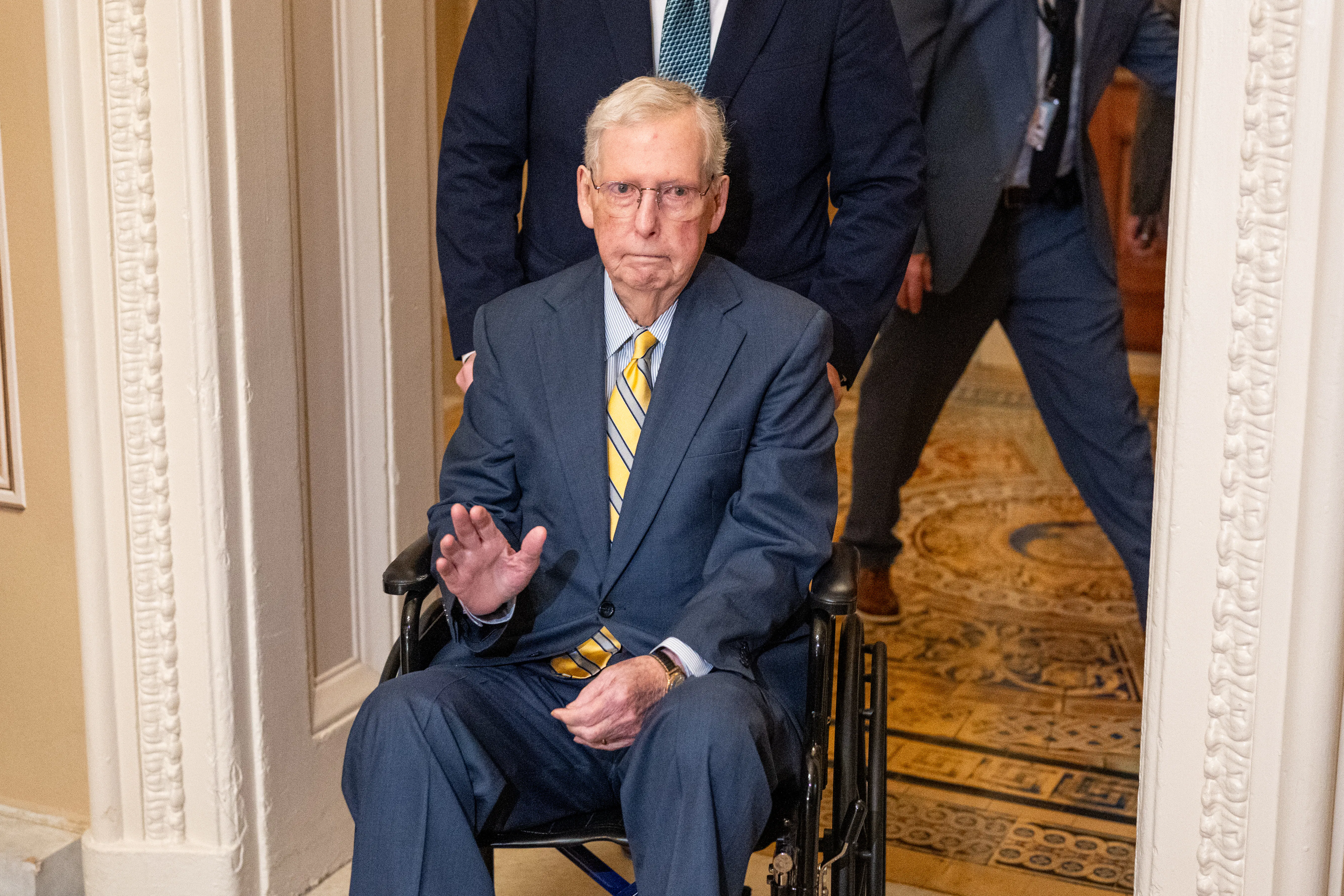 Mitch McConnell waves while being pushed in a wheelchair at the Capitol.