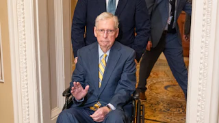 Mitch McConnell waves while being pushed in a wheelchair at the Capitol.
