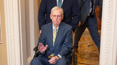 Mitch McConnell waves while being pushed in a wheelchair at the Capitol.