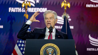 Dr. Mehmet Oz stands behind a podium with the presidential seal on it, wearing a dark suit and red tie, while raising his right hand. Behind him, a backdrop reads "Faith and Freedom" while an American flag is visible next to another flag.