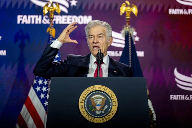 Dr. Mehmet Oz stands behind a podium with the presidential seal on it, wearing a dark suit and red tie, while raising his right hand. Behind him, a backdrop reads "Faith and Freedom" while an American flag is visible next to another flag.