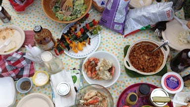 Dishes of crab, baked beans, vegetables on a skewer, salad, and condiments sit on a table alongside plastic cutlery and paper plates.