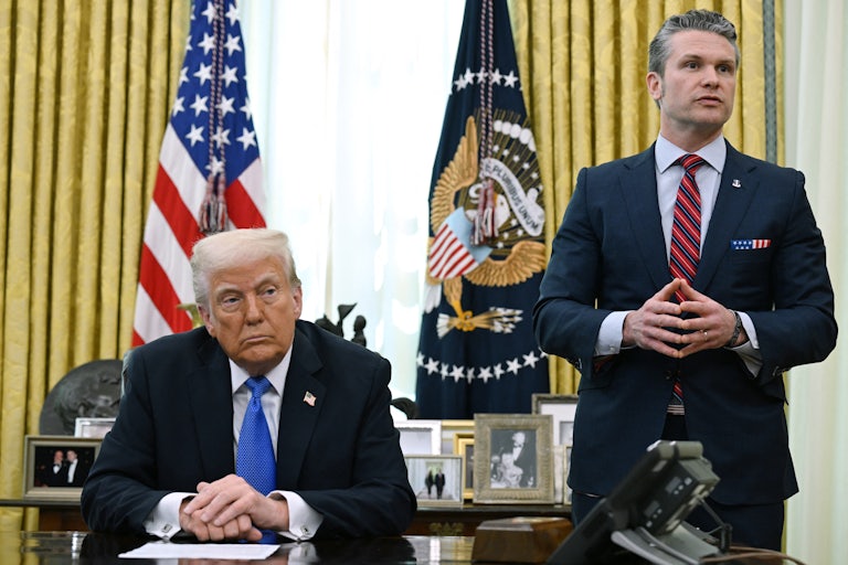 Donald Trump clasps his hands on his desk in the White House as Defense Secretary Pete Hegseth stands next to him speaking (presumably to reporters).