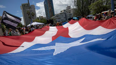 Demonstrators in San Juan, Puerto Rico hold flat a big flag of Puerto Rico, with other smaller flags of Puerto Rico visible in the crowd. Behind the demonstrators, trees and tall buildings are visible.