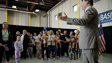 A group of white people, including children, stand waving U.S. flags as Deputy Secretary of State Christopher Landau greets them.
