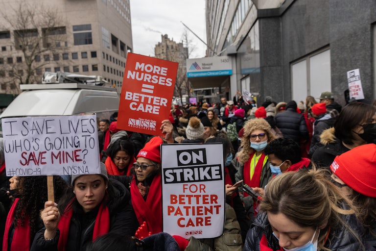 Striking health care workers hold up signs like "I SAVE LIVES WHO'S GOING TO SAVE MINE?," "ON STRIKE FOR BETTER CARE," and "MORE NURSES = BETTER CARE."