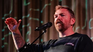 Graham Platner speaks into a microphone holding his hand up