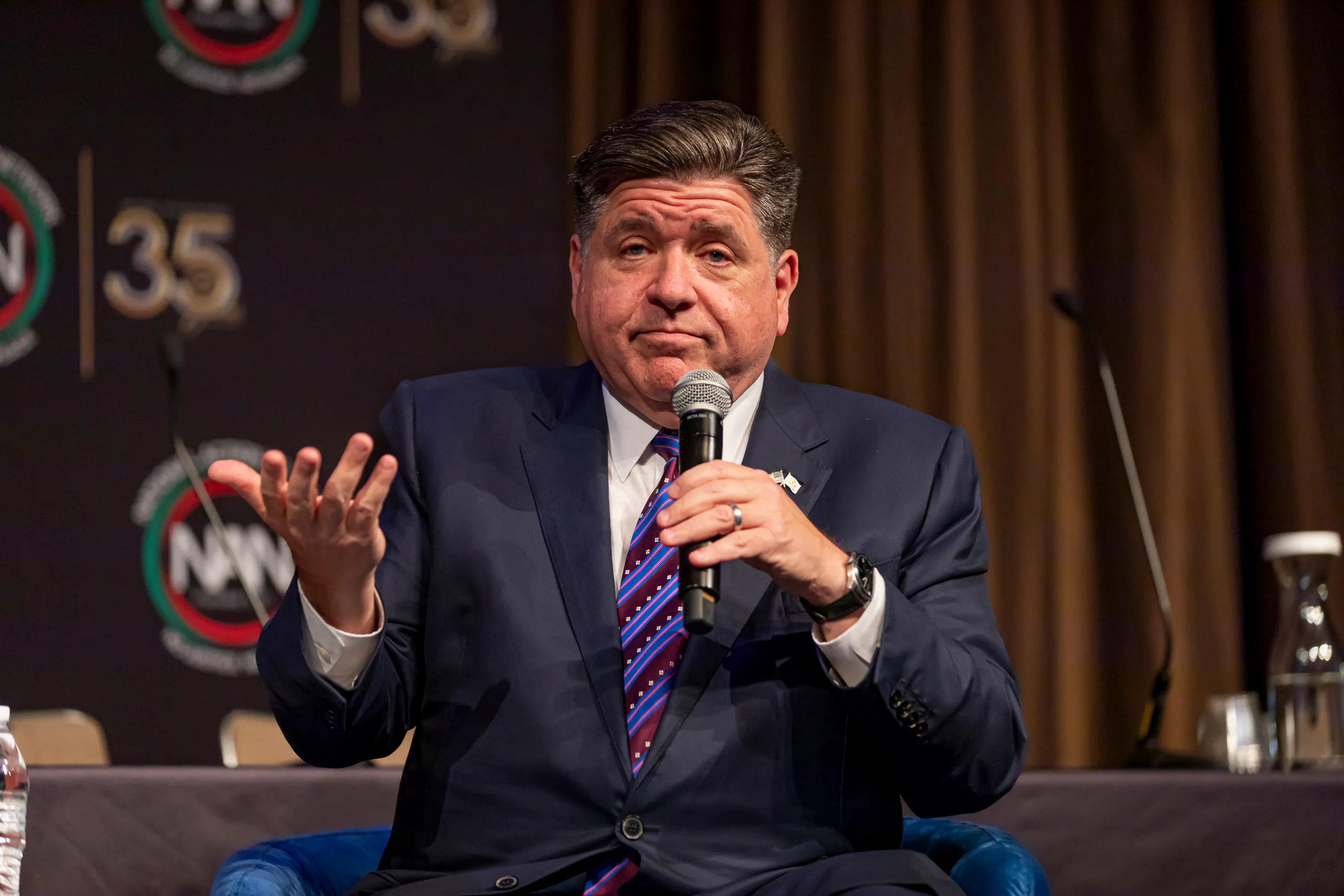 JB Pritzker sits wearing a navy blue suit and a blue striped tie, holding a microphone in one hand and having his other hand in a palms-up position. 