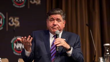 JB Pritzker sits wearing a navy blue suit and a blue striped tie, holding a microphone in one hand and having his other hand in a palms-up position.