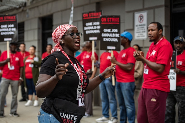 Striking workers wearing red hold up signs. A woman in the front speaks to them animatedly. They are all Black.