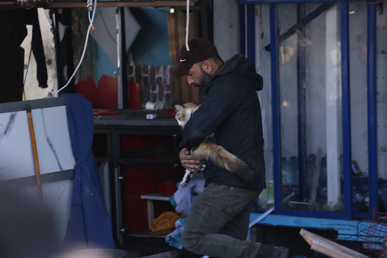 A man carries a cat in his arms among rubble.