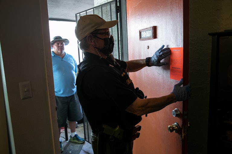 A law enforcement officer posts an eviction notice on the painted red door of an apartment while another man looks on