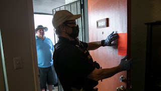 A law enforcement officer posts an eviction notice on the painted red door of an apartment while another man looks on