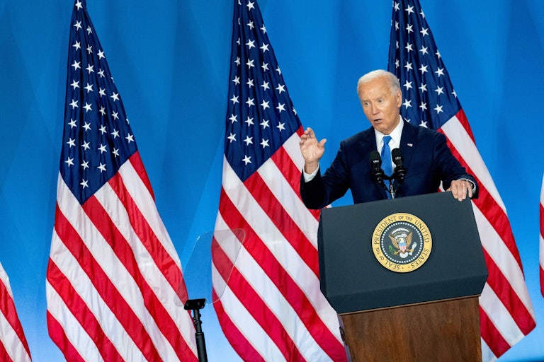 Joe Biden speaking at a lectern and making a hand gesture. A row of U.S. flags are behind him.