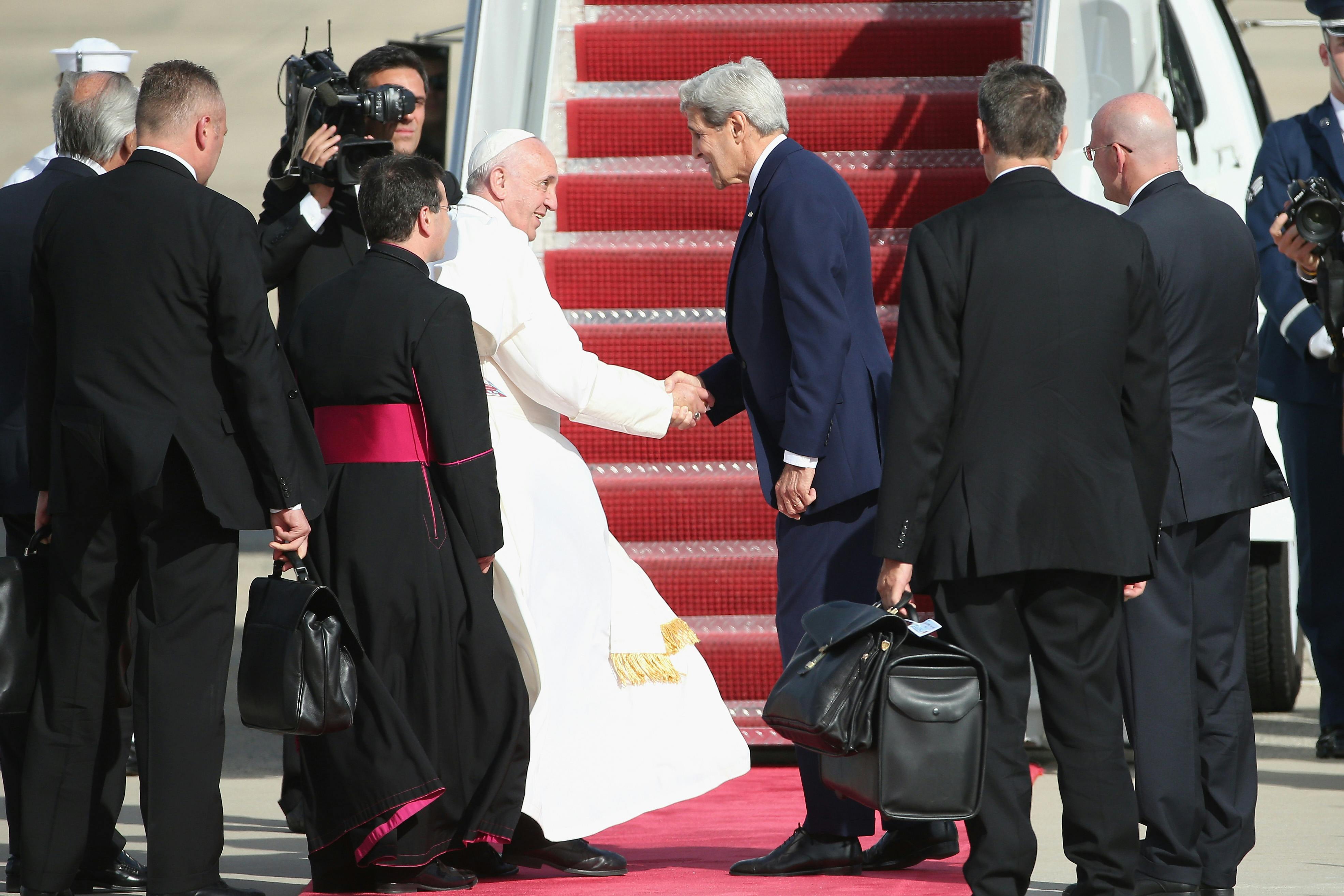 Pope Francis shakes hands with John Kerry on a tarmac, surrounded by aides.