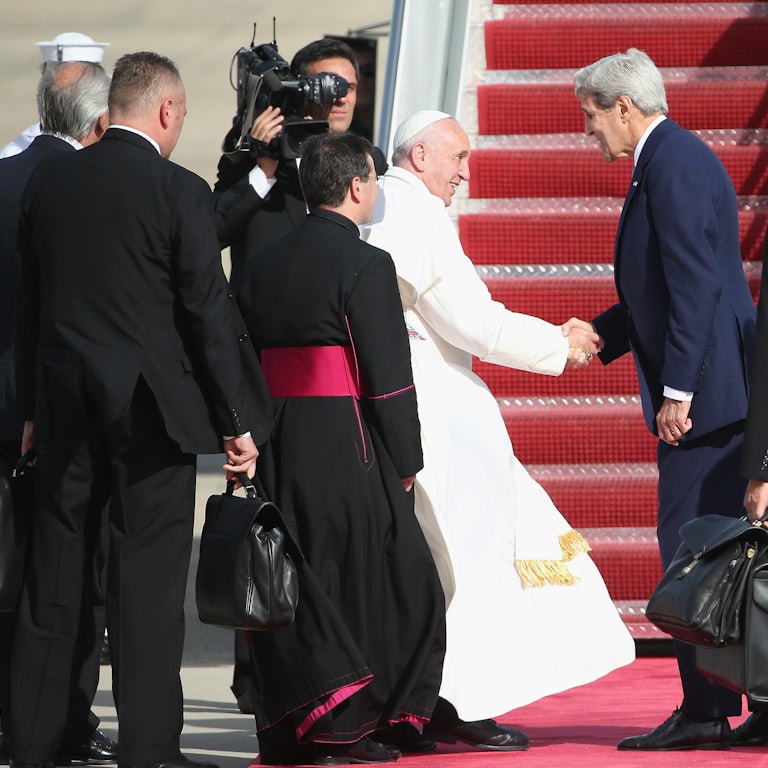 Pope Francis shakes hands with John Kerry on a tarmac, surrounded by aides.