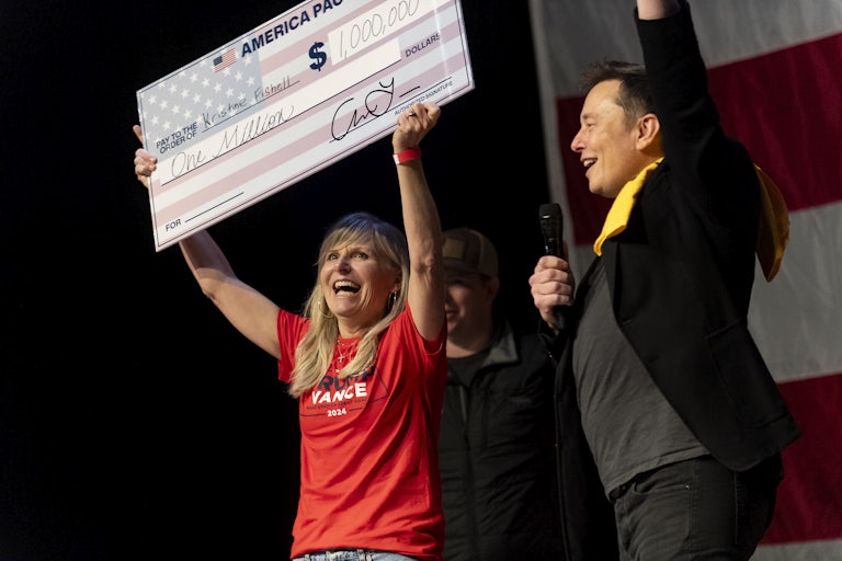 Elon Musk raises a hand in the air and smiles as a woman next to him on the stage holds a giant $1 million check in the air with both hands and smiles