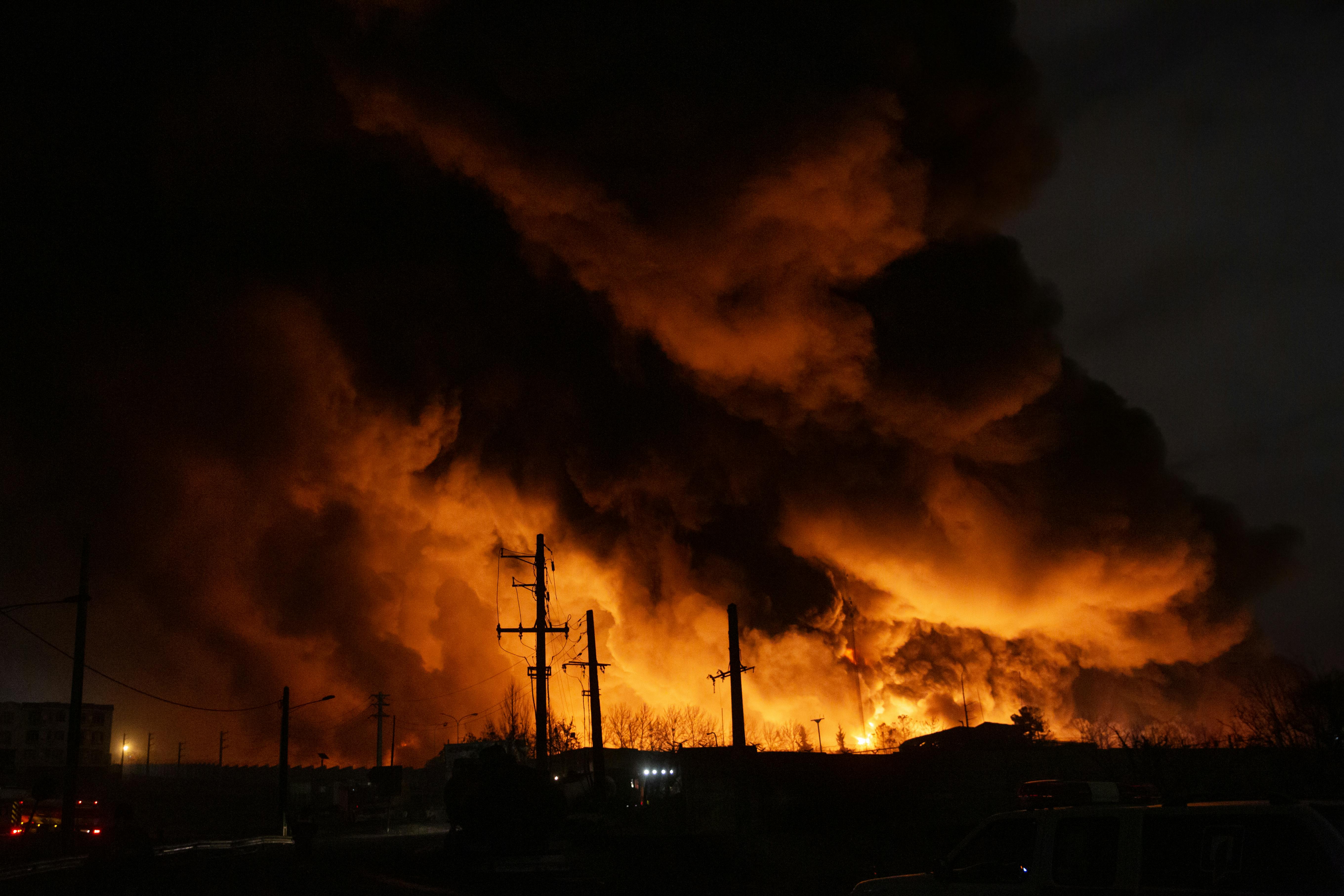 Clouds of black smoke, lit up with orange light from fires, rise up over a dark landscape at night.