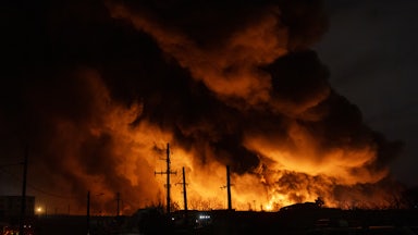 Clouds of black smoke, lit up with orange light from fires, rise up over a dark landscape at night.