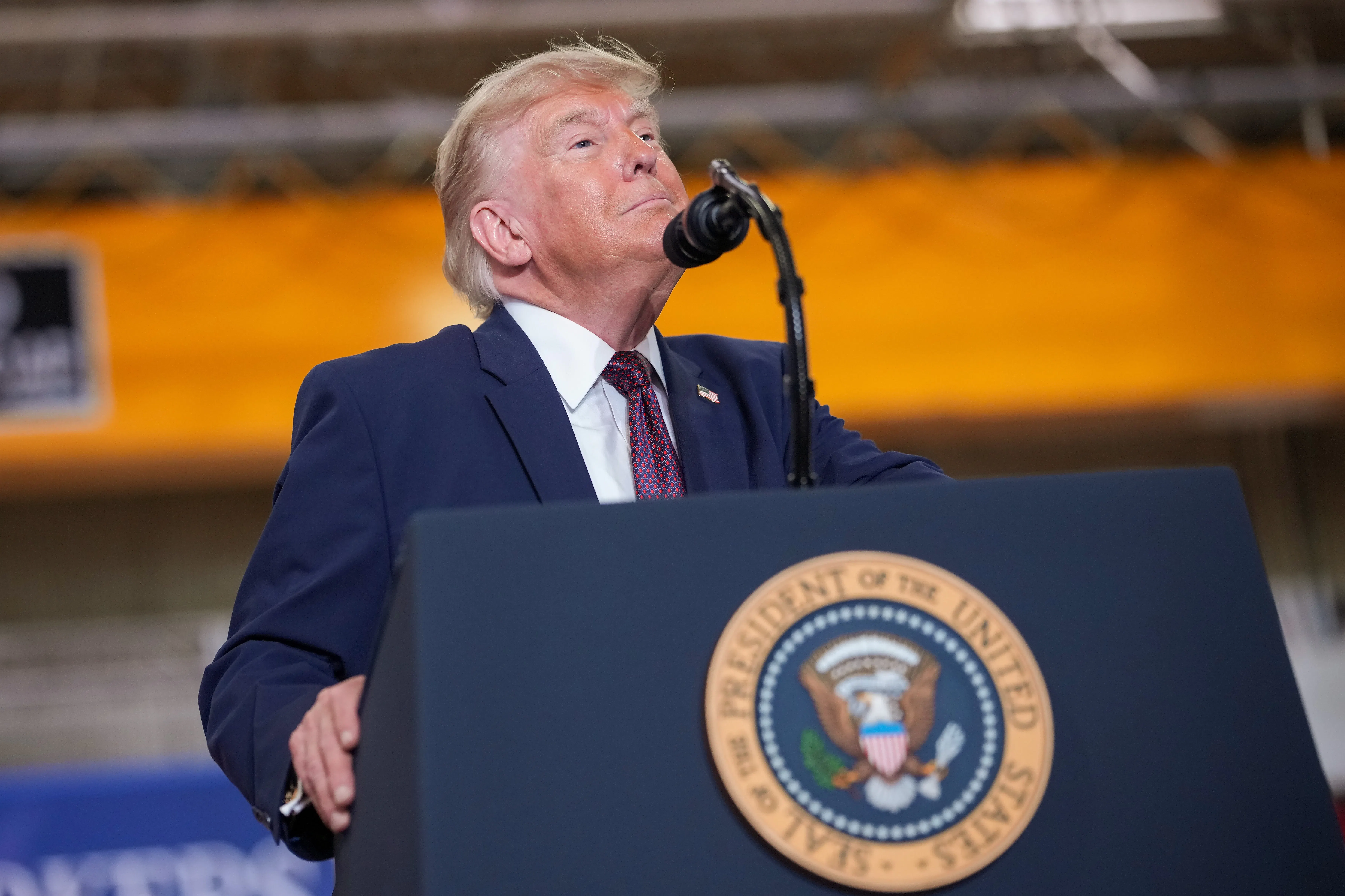 Trump, wearing a navy blue suit with a dark tie, sticks his chin up while standing behind a microphone and podium with the presidential seal. 