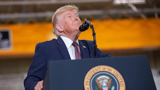 Trump, wearing a navy blue suit with a dark tie, sticks his chin up while standing behind a microphone and podium with the presidential seal.