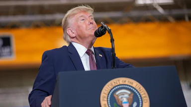Trump, wearing a navy blue suit with a dark tie, sticks his chin up while standing behind a microphone and podium with the presidential seal.