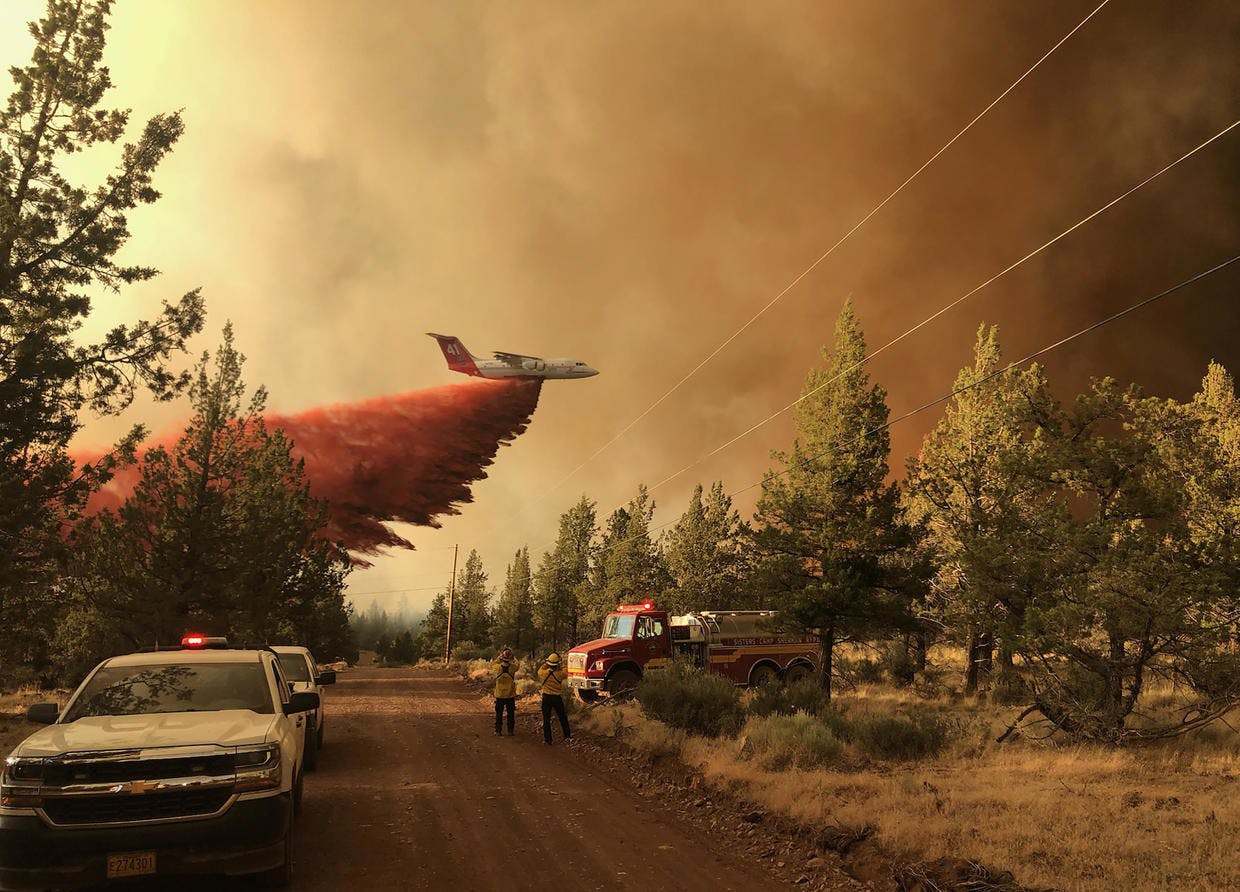 An aircraft dumps fire retardant as emergency vehicles stand in the foreground.