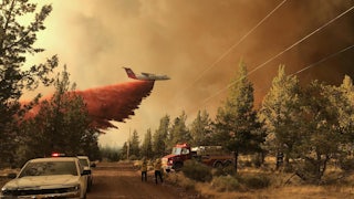 An aircraft dumps fire retardant as emergency vehicles stand in the foreground.