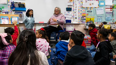 Kids sit on the floor and listen to their teacher at the front of the room