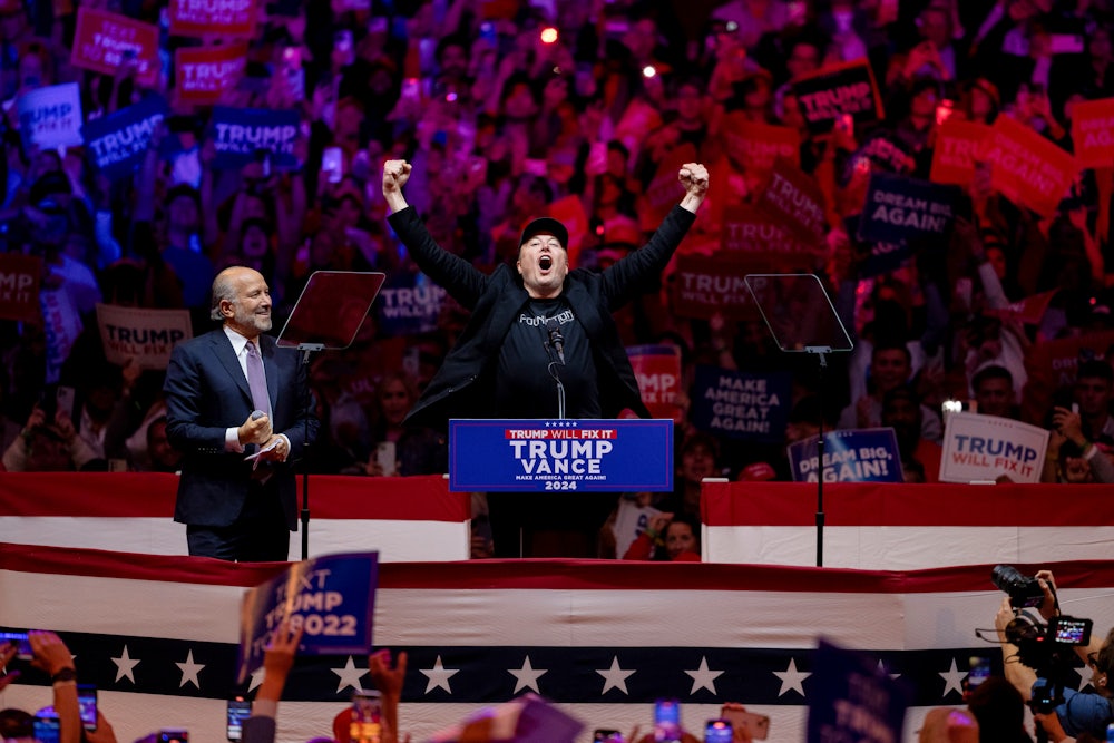Musk raises both fists above his head triumphantly at a podium while Lutnik looks on and supporters hold signs in the background.