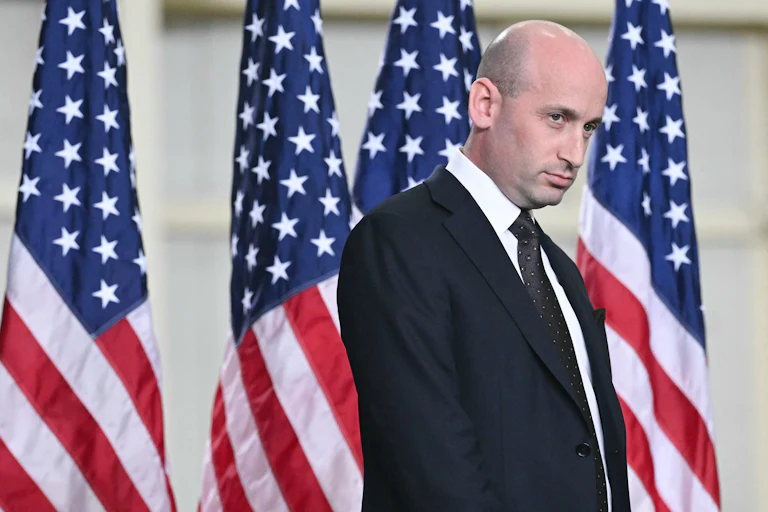 Stephen Miller glares as he stands in front of a row of U.S. flags.